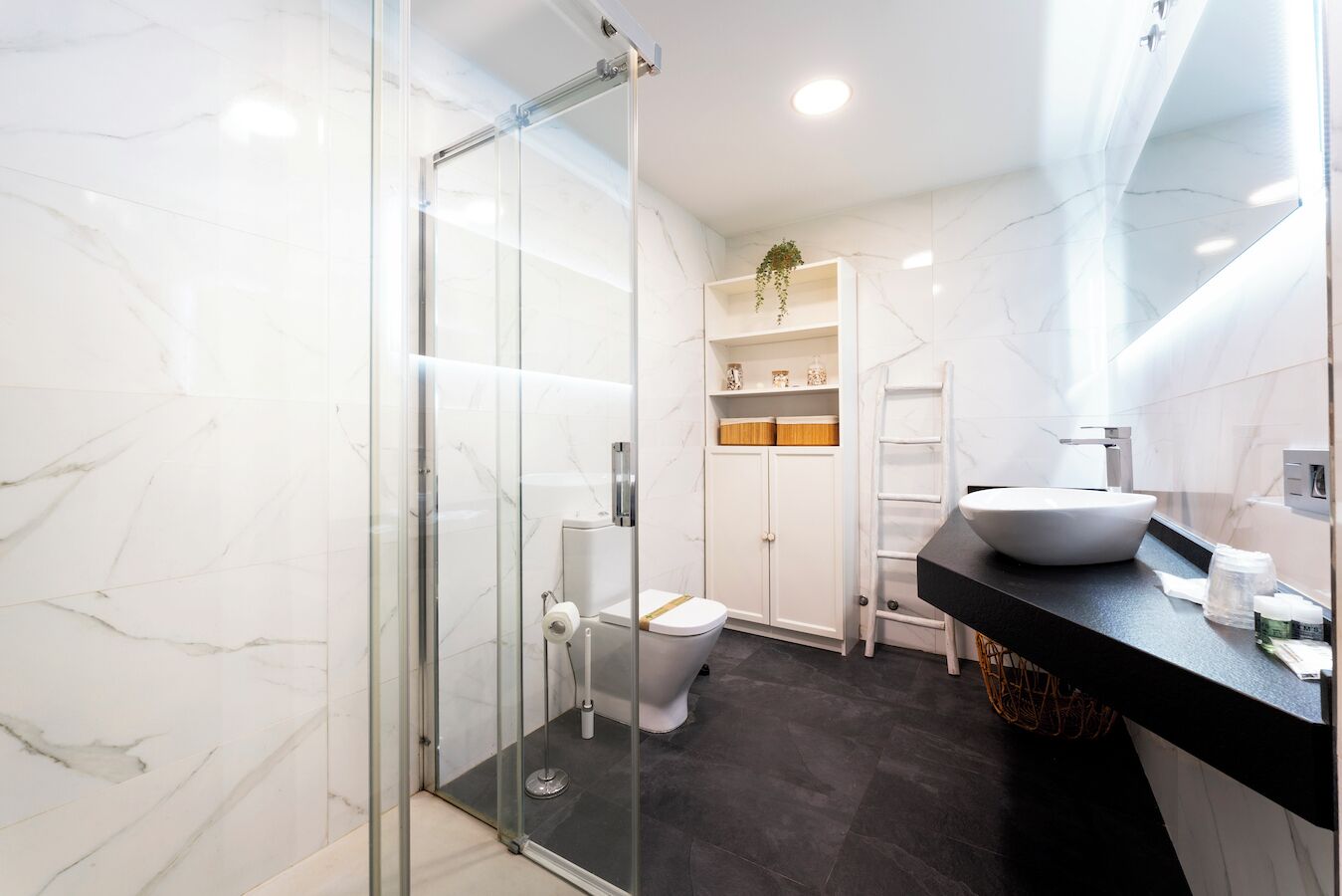 A modern bathroom with a glass shower enclosure, a white toilet, a black countertop with a vessel sink, and a white cabinet with shelves.