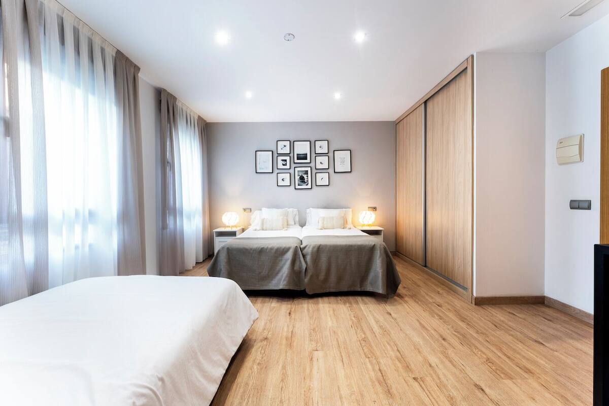 A hotel room with two beds, a wooden floor, a wall with framed pictures, and a large window with curtains.