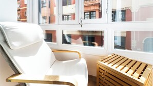 Een kamer met een witte stoel, een houten kist en een raam met uitzicht op gebouwen.