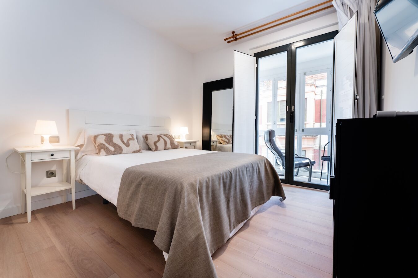 Un dormitorio con cama, mesitas de noche, televisor y un balcón con una silla.