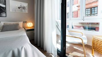 Una habitación de hotel moderna con una cama, una mesita de noche con lámpara, una silla y vistas a edificios a través de un ventanal.