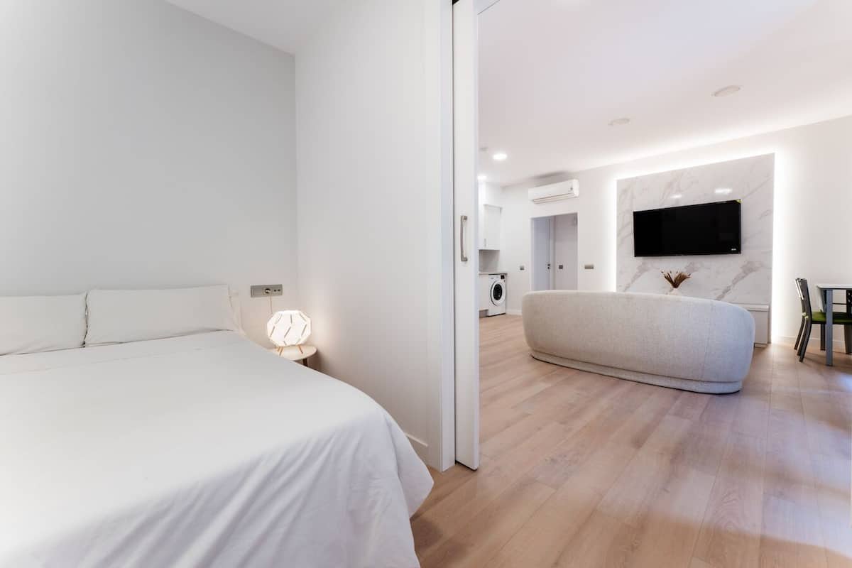 A modern hotel room with a large bed, a seating area, a flat-screen TV, and a washing machine in the background.