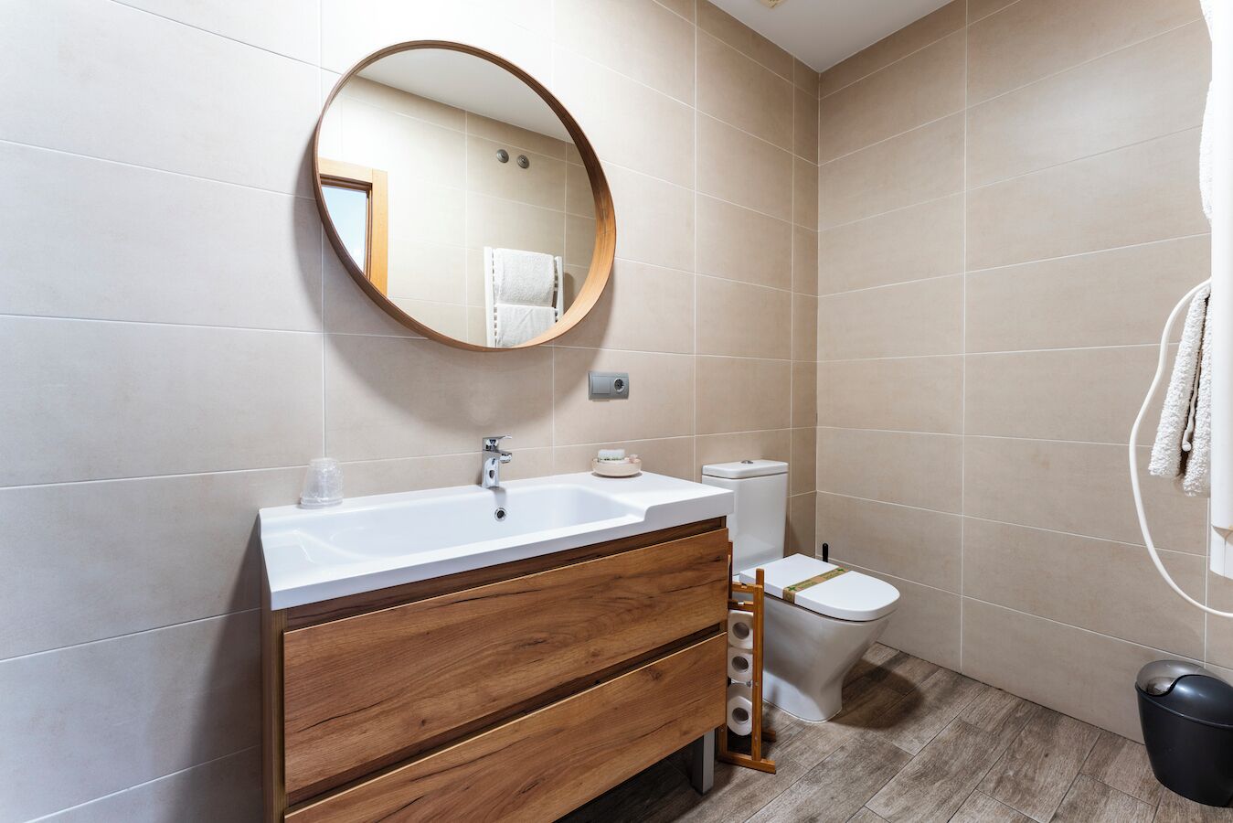 A bathroom with a wooden vanity, a round mirror, a toilet, and a trash bin.