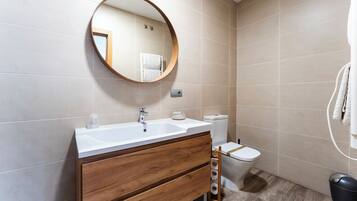 Une salle de bain avec un meuble-lavabo en bois, un miroir rond, des toilettes et une poubelle.