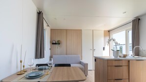 A modern kitchen with wooden cabinets, a dining area with a table set for two, and a view through a window. - Caravan Bredene on the Coast (Bredene)