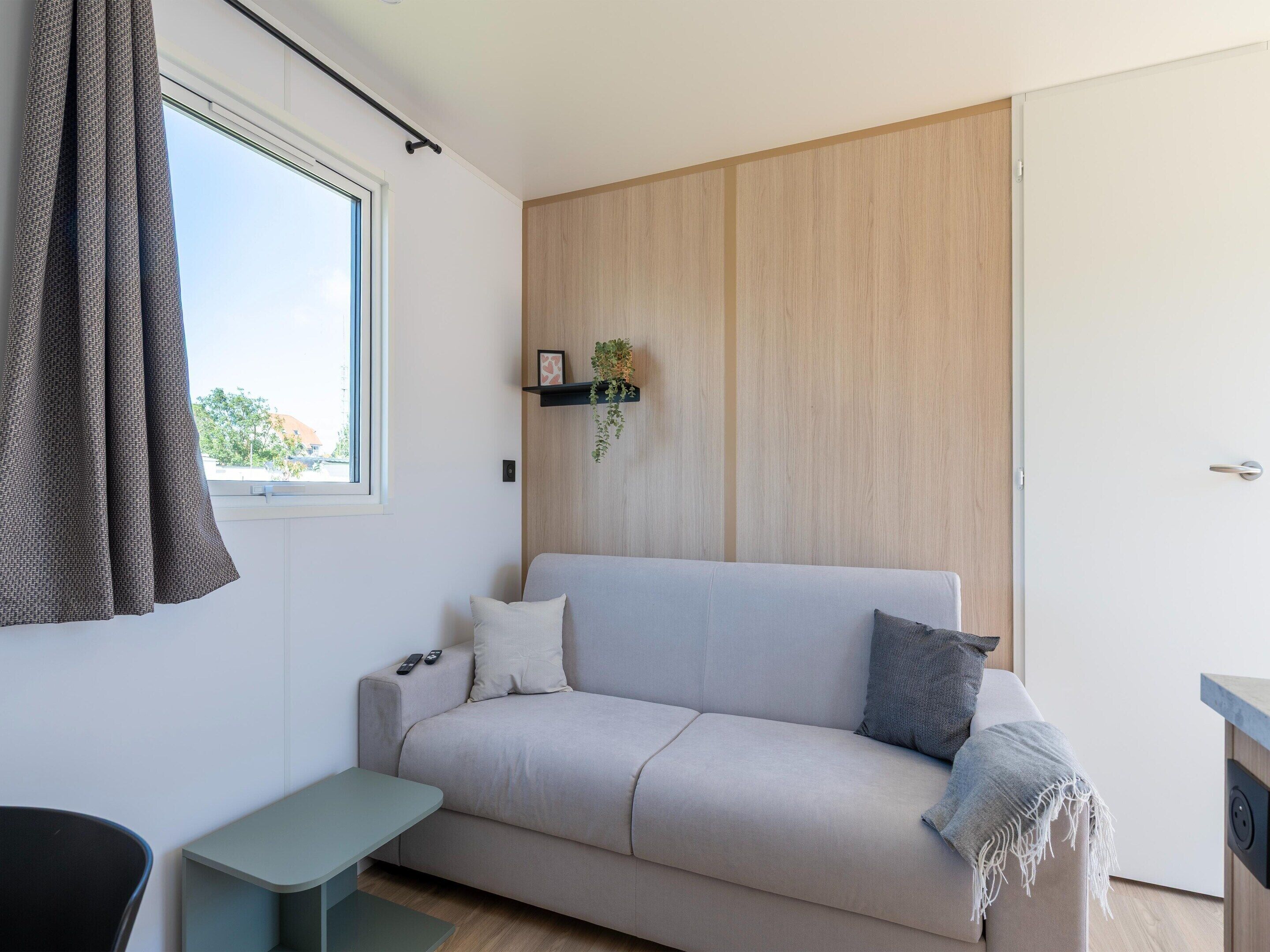 Un salon moderne comprenant un canapé, une petite table, une fenêtre avec des rideaux, un mur recouvert de panneaux en bois et une porte.