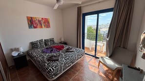 Ein Schlafzimmer mit Bett, Nachttisch, Schreibtisch, Stuhl, Balkon mit Aussicht und Deckenventilator.