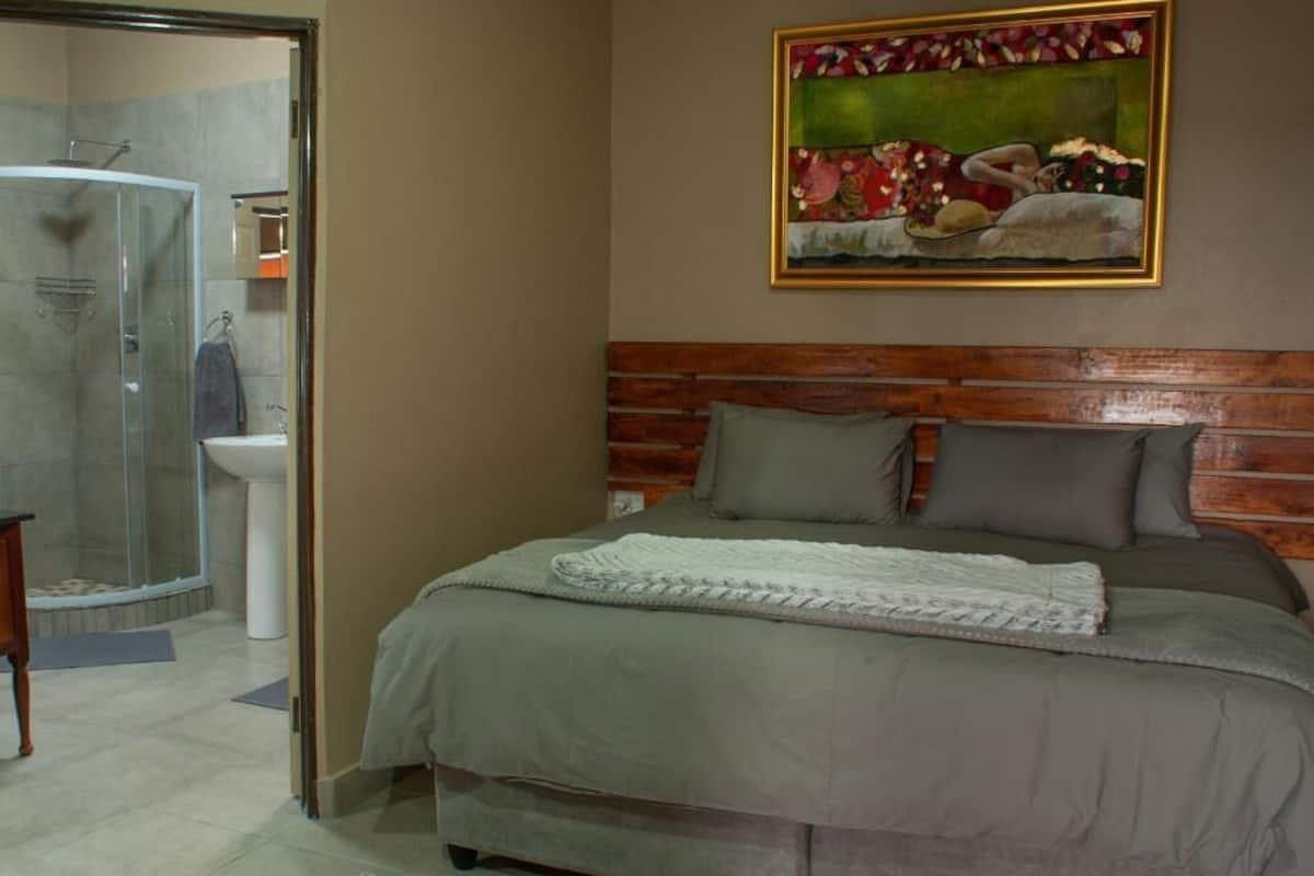 Une chambre avec une tête de lit en bois, un lit recouvert d’une couverture grise, un tableau accroché au mur et un espace douche.