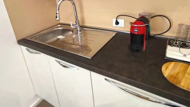 Una cucina con lavello in acciaio inossidabile, mobili bianchi, una macchina per caffè e un tagliere di legno.