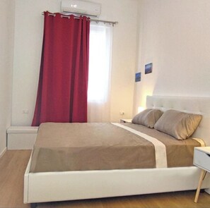 Ein Schlafzimmer mit Bett, Nachttisch, Wand-Klimaanlage und einem Fenster mit rotem Vorhang.