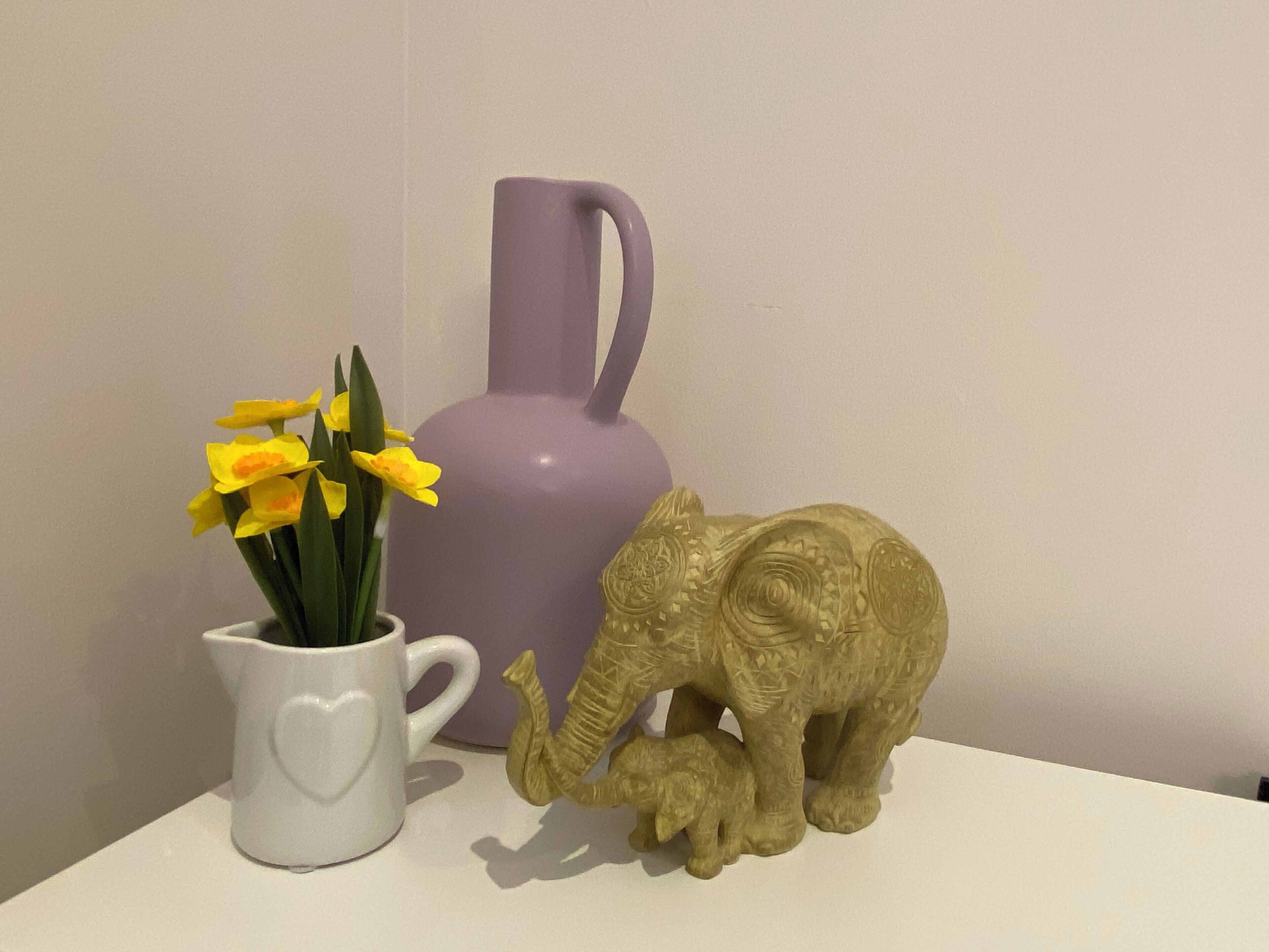 A white mug with a heart design, a purple vase, and a golden elephant figurine on a white surface.