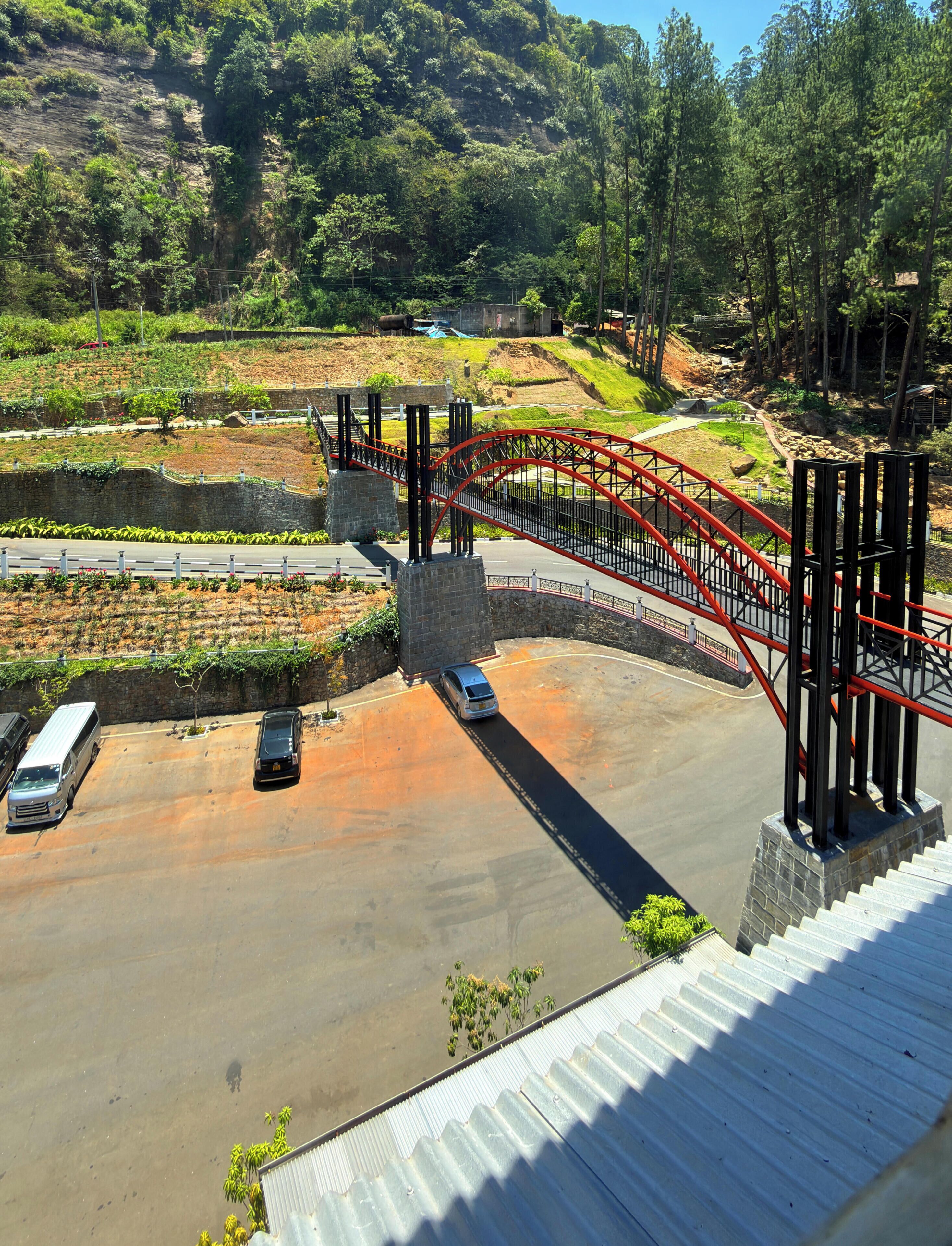 A parking area with a red bridge, vehicles, and a mountainous backdrop.