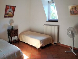 A bedroom with a bed, a radiator, a window, a standing fan, and a small table with a lamp.