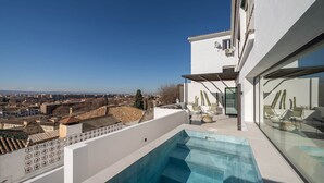 Une piscine sur le toit offrant une vue sur la ville, un salon aménagé dans une structure entièrement vitrée et un bâtiment blanc surmonté d’une antenne parabolique.