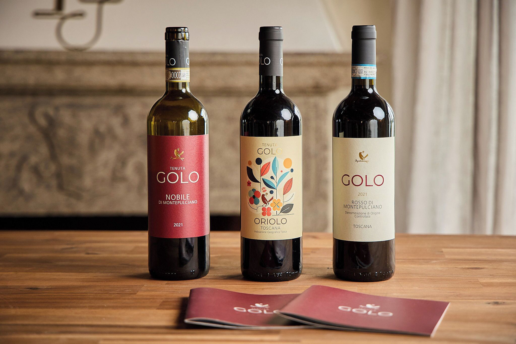 Tre flasker Tenuta Golo vin på et træbord med en rød brochure ved siden af.