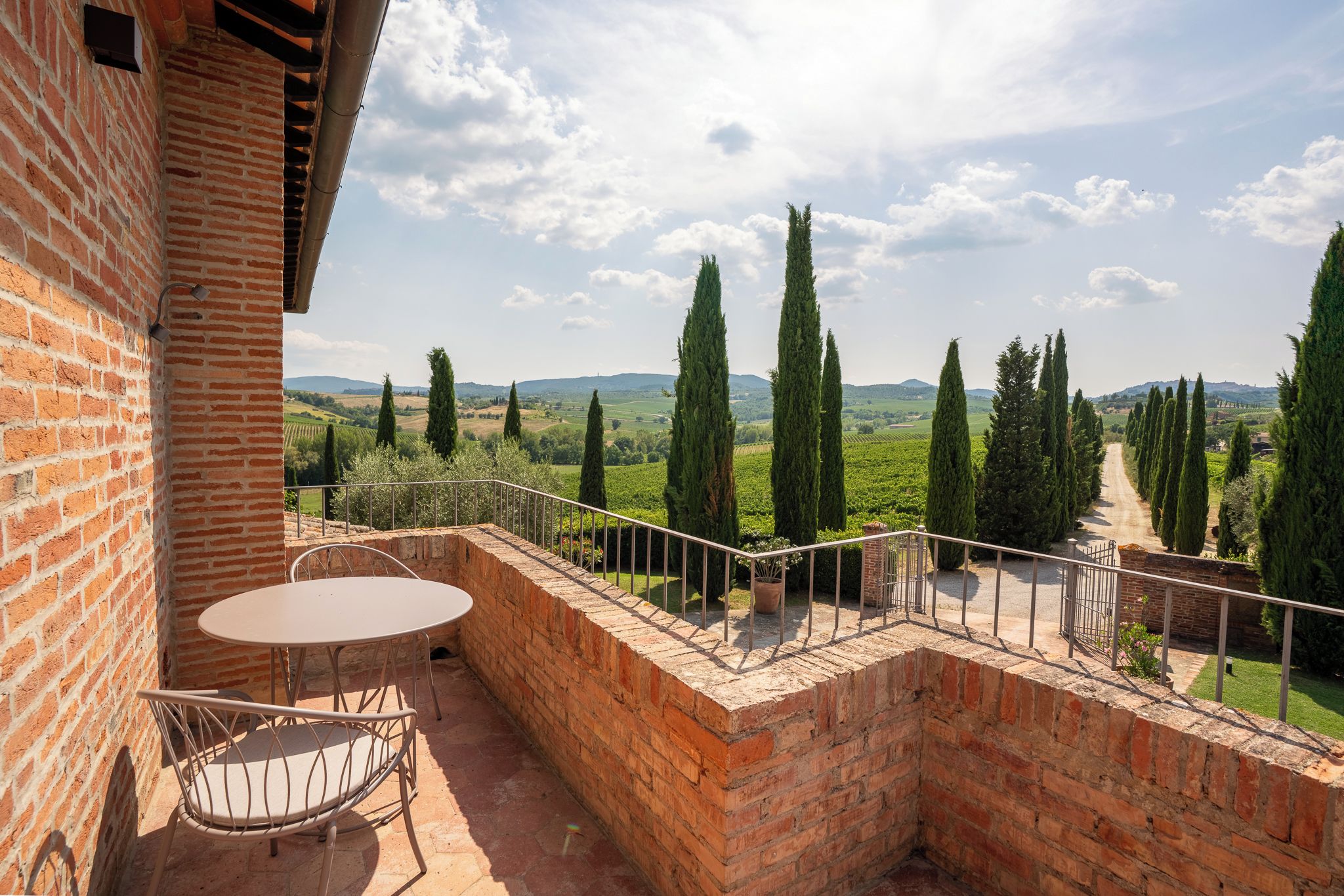 A brick balcony with a table and chairs, overlooking a vineyard with tall cypress trees.