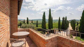 A brick balcony with a table and chairs, overlooking a vineyard with tall cypress trees.