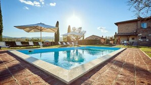A swimming pool with lounge chairs and umbrellas, surrounded by a brick patio and a house. - Tenuta Golo (Montepulciano)
