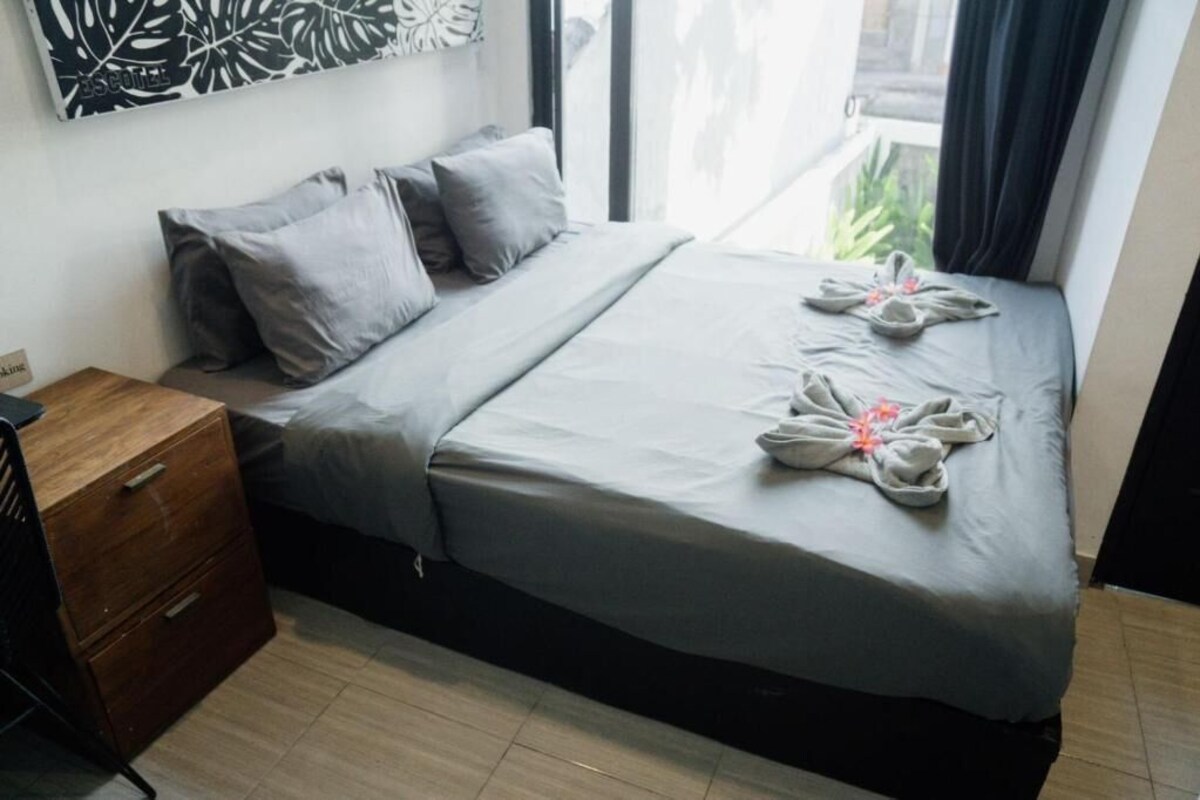 A neatly made bed with a grey bedspread and pillows, a wooden nightstand with drawers, and a wall art piece above the bed.