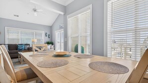 A dining area with a wooden table, woven placemats, and a view of the city through large windows with blinds. - Chonky Waves by Brunswick VR: 4BR Condo w/ Ocean Views & Resort Amenities (Ocean Isle Beach)