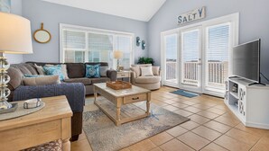 A living room with a sofa, coffee table, TV stand, and a ceiling fan. - Chonky Waves by Brunswick VR: 4BR Condo w/ Ocean Views & Resort Amenities (Ocean Isle Beach)