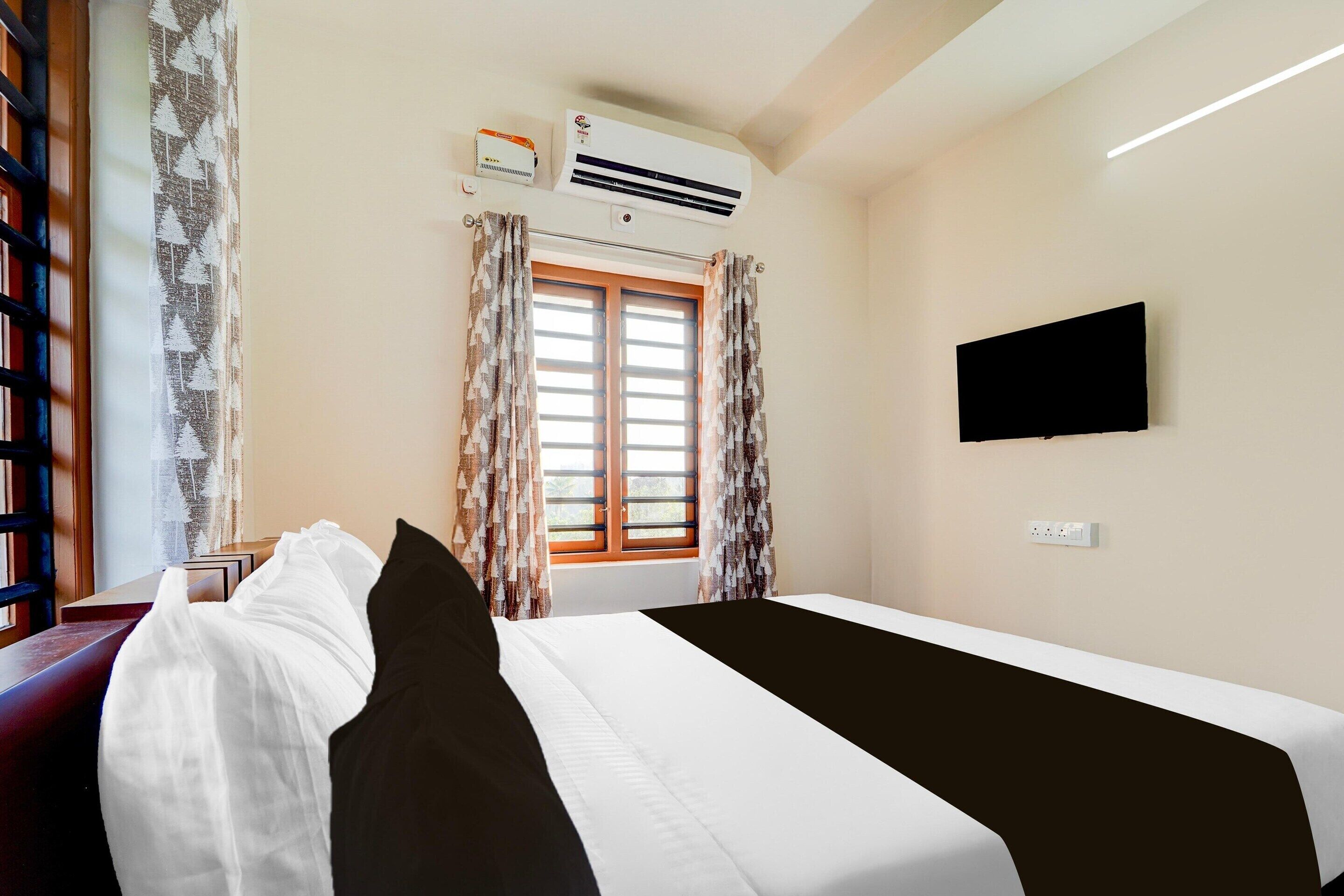 A hotel room with a bed, a flat-screen TV, an air conditioning unit, and a window with curtains.