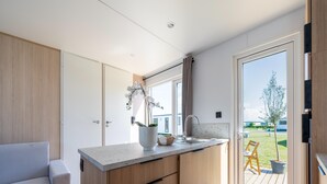 A modern kitchen with a built-in island, a dining area with a light-colored sofa, and a sliding glass door leading to an outdoor deck. - Caravan Bredene by the sea (Bredene)