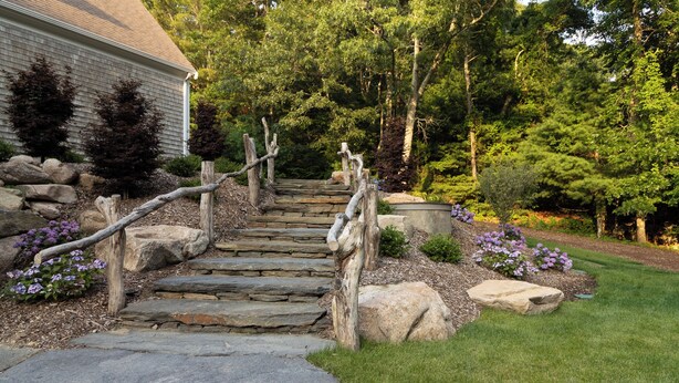 Eine Steintreppe führt zu einem Haus mit einem hölzernen Geländer, umgeben von einem gepflegten Garten mit verschiedenen Sträuchern und Blumen.