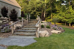 A stone staircase leading to a house with a wooden handrail, surrounded by a well-maintained garden with various shrubs and flowers. - Newly remodeled home on Cape Cod by Old Silver Beach (Falmouth)