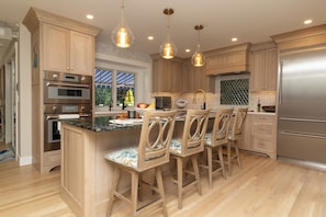 A modern kitchen with wooden cabinets, stainless steel appliances, a central island with bar stools, pendant lighting, and a view of the outdoors. - No crowds after Labor Day Week on Cape Cod  at Old Silver Beach (Falmouth)