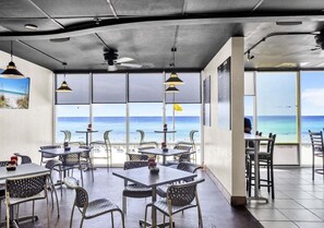 A restaurant with outdoor seating overlooking the beach, featuring metal chairs and tables, large windows, and a ceiling fan. - Coastal Calm 1411-Gulf Views-Sleeps 8 (Panama City Beach)