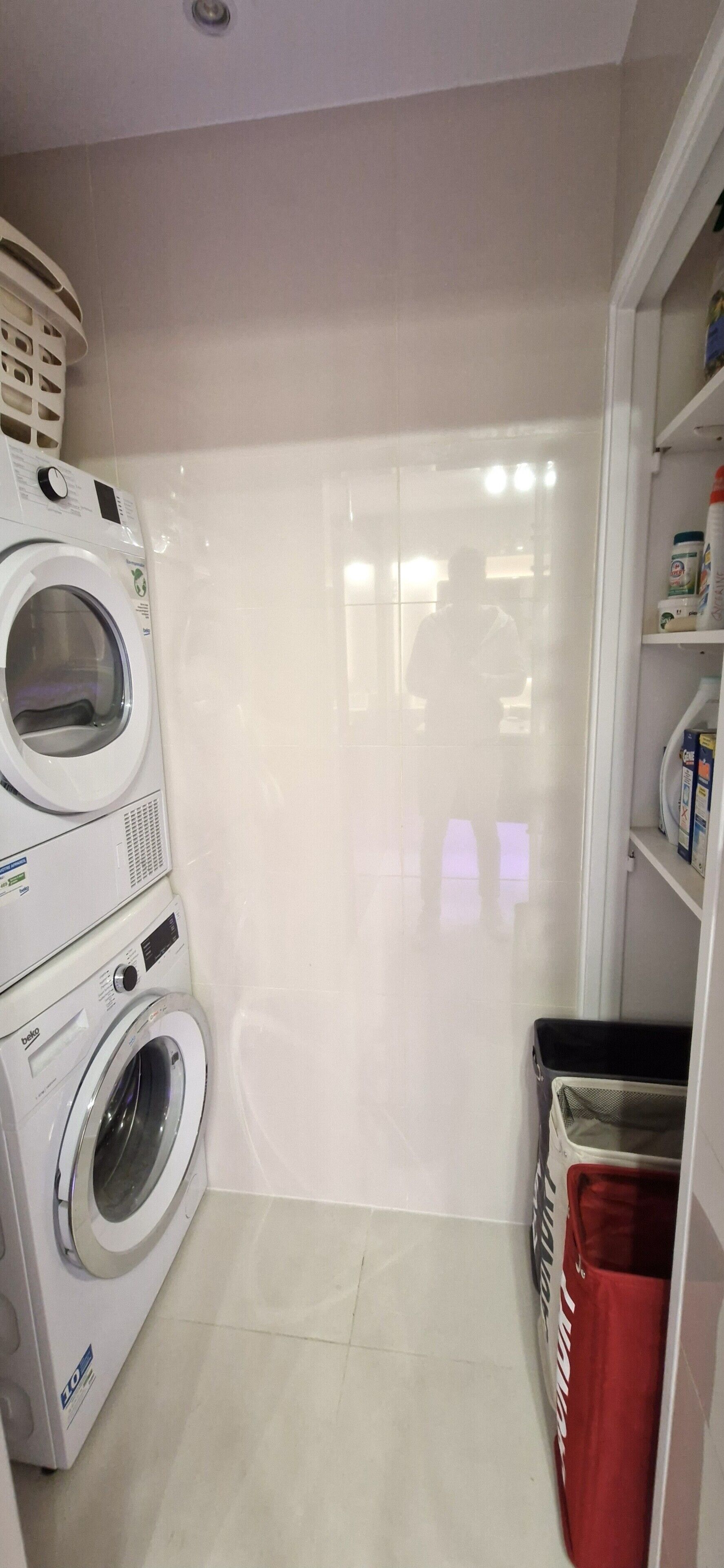 A laundry room with stacked washing machines, a red laundry basket, and a shelf with cleaning supplies.