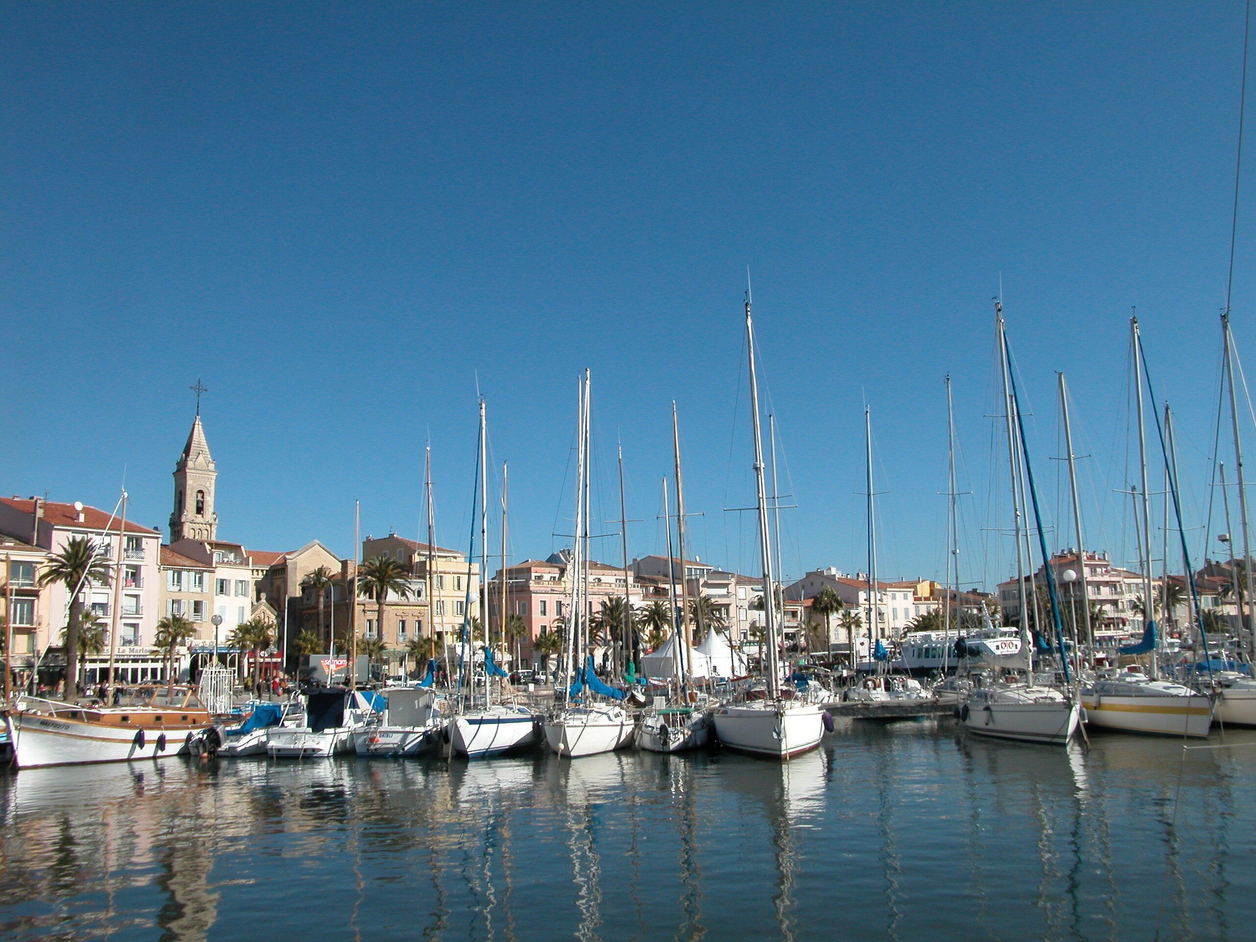 A marina with multiple sailboats docked, a prominent church tower, and residential buildings in the background.