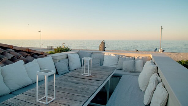 Un área de descanso junto a la playa con una mesa de madera, cojines blancos y vista despejada al mar.