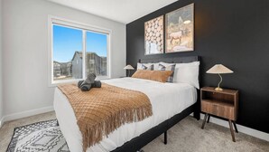 A bedroom with a large bed, a nightstand, a lamp, a window with a view, and artwork on the wall. - Luxurious 4BR Home | Near Banff | YYC | Garage (Calgary)