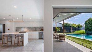 A modern kitchen with a central island, bar stools, and a view of a swimming pool and backyard. - Mooloolaba Canal Retreat with Pool, Sleeps 12 (Mooloolaba)