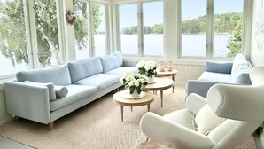 A room with a light blue sofa, a white armchair, and a small round table with a vase of flowers. Large windows provide a view of a lake and trees. - Waterfront Retreat - Frontline Oslofjord | Small boat included | Fireplace (Akershus)