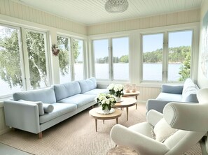 Ein Zimmer mit einem hellblauen Sofa, einem weißen Sessel und einem kleinen runden Tisch mit einem Blumenstrauß. Große Fenster bieten Blick auf einen See und Bäume.