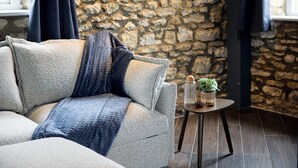 Una habitación acogedora con pared de piedra, un sofá gris con una manta azul y una mesita con una planta y dos vasos.
