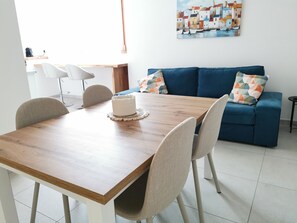 A dining area with a wooden table, four chairs, a white cake, a blue sofa with patterned cushions, and a painting of a coastal scene. - Domus Soli - Sunny Elegance and Contemporary Design in Teulada (Teulada)