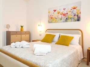 A neatly made bed with white and yellow pillows, a bedside table with a vase, and a wall painting of flowers. - Domus Soli - Sunny Elegance and Contemporary Design in Teulada (Teulada)