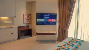 Et moderne hotelværelse med et fladskærms-tv der viser en velkomstskærm, et lille skrivebord, et siddeområde og en seng med et sengetæppe med et geometrisk mønster.