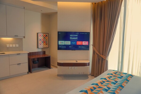 A modern hotel room with a flat-screen TV displaying a welcome screen, a small desk, a seating area, and a bed with a geometric-patterned bedspread.