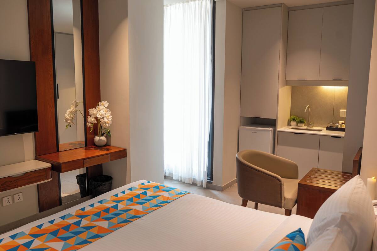 A hotel room with a bed, a desk with a TV, a chair, a small kitchen area, and a bathroom.
