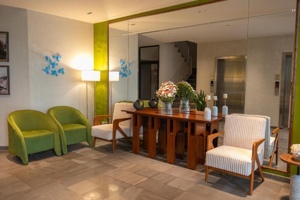 A hotel lobby with a wooden reception desk, two green armchairs, a white armchair, and a mirror reflecting the elevator.