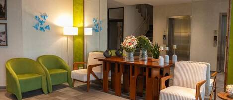 Un hall de hotel con un mostrador de recepción de madera, dos sillones verdes, un sillón blanco y un espejo que refleja los ascensores.