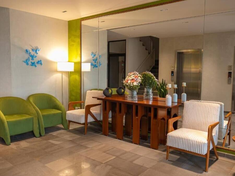 A hotel lobby with a wooden reception desk, two green armchairs, a white armchair, and a mirror reflecting the elevator.
