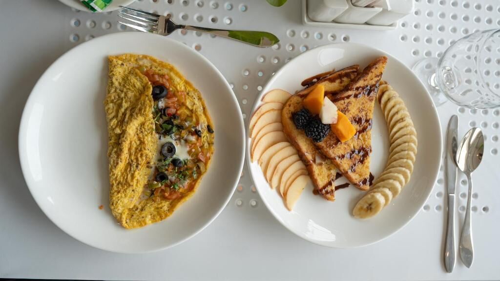 Dos platos de comida, uno con una tortilla y otro con tortitas, sobre una mesa blanca con un vaso y cubiertos.