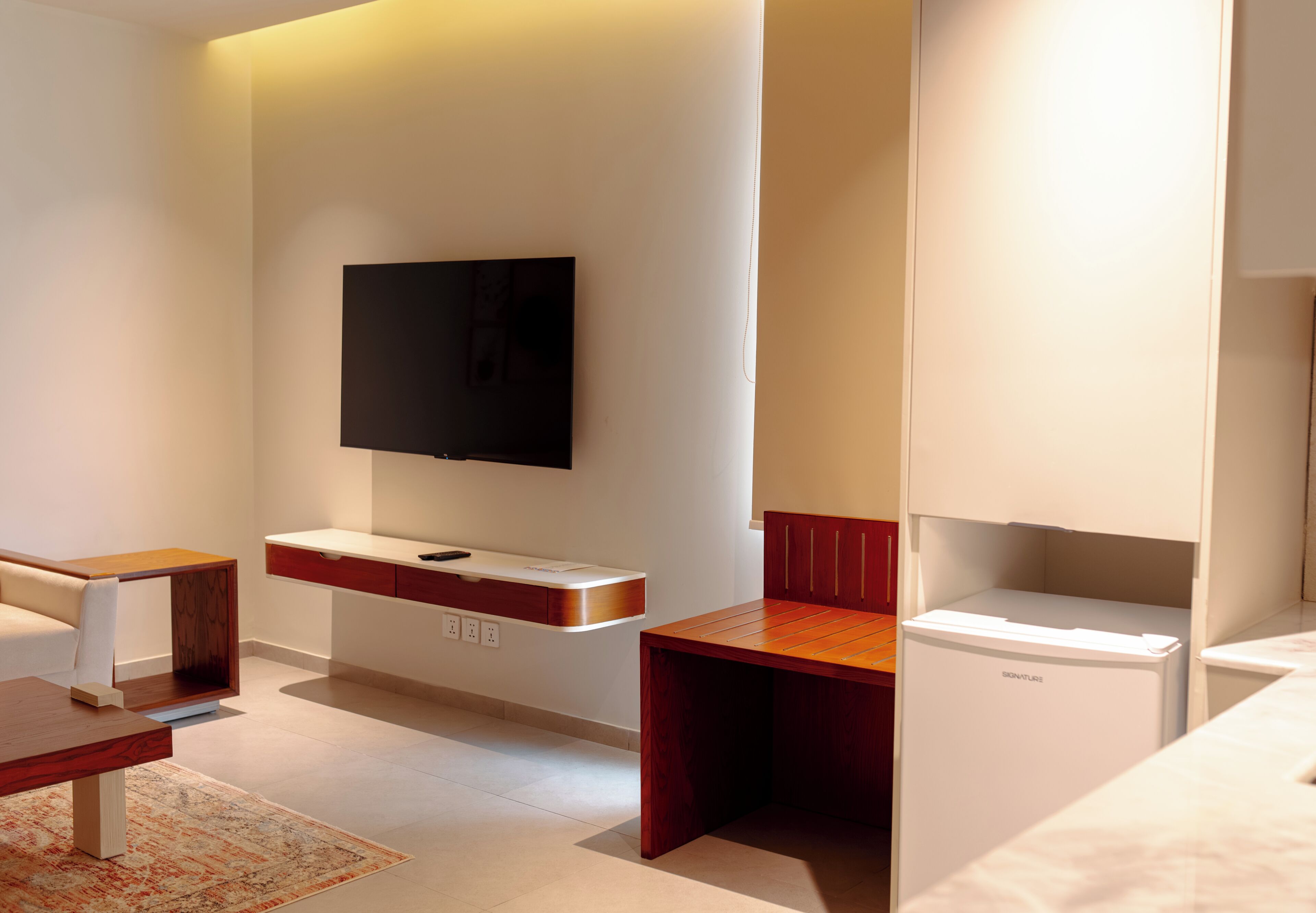 A modern hotel room with a flat-screen TV mounted on the wall, a wooden desk, a small refrigerator, and a sofa.