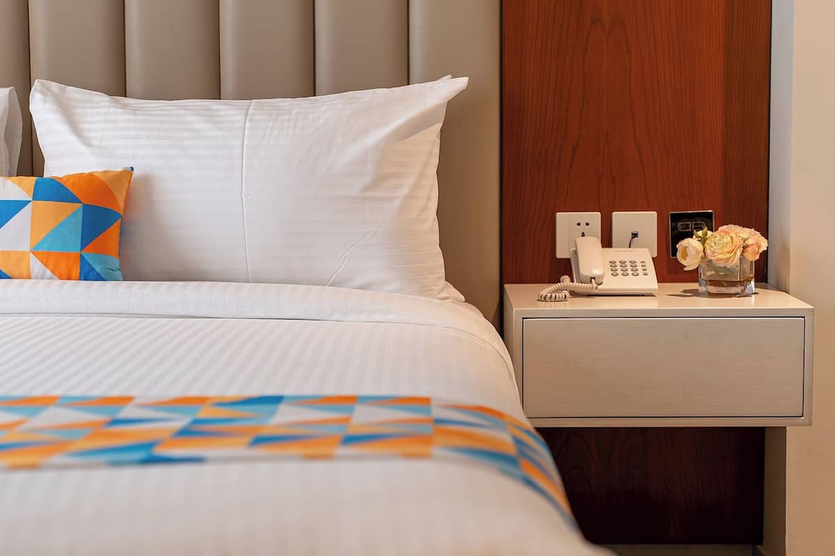A hotel room with a bed, a nightstand with a phone and a vase of flowers, and a wooden headboard.